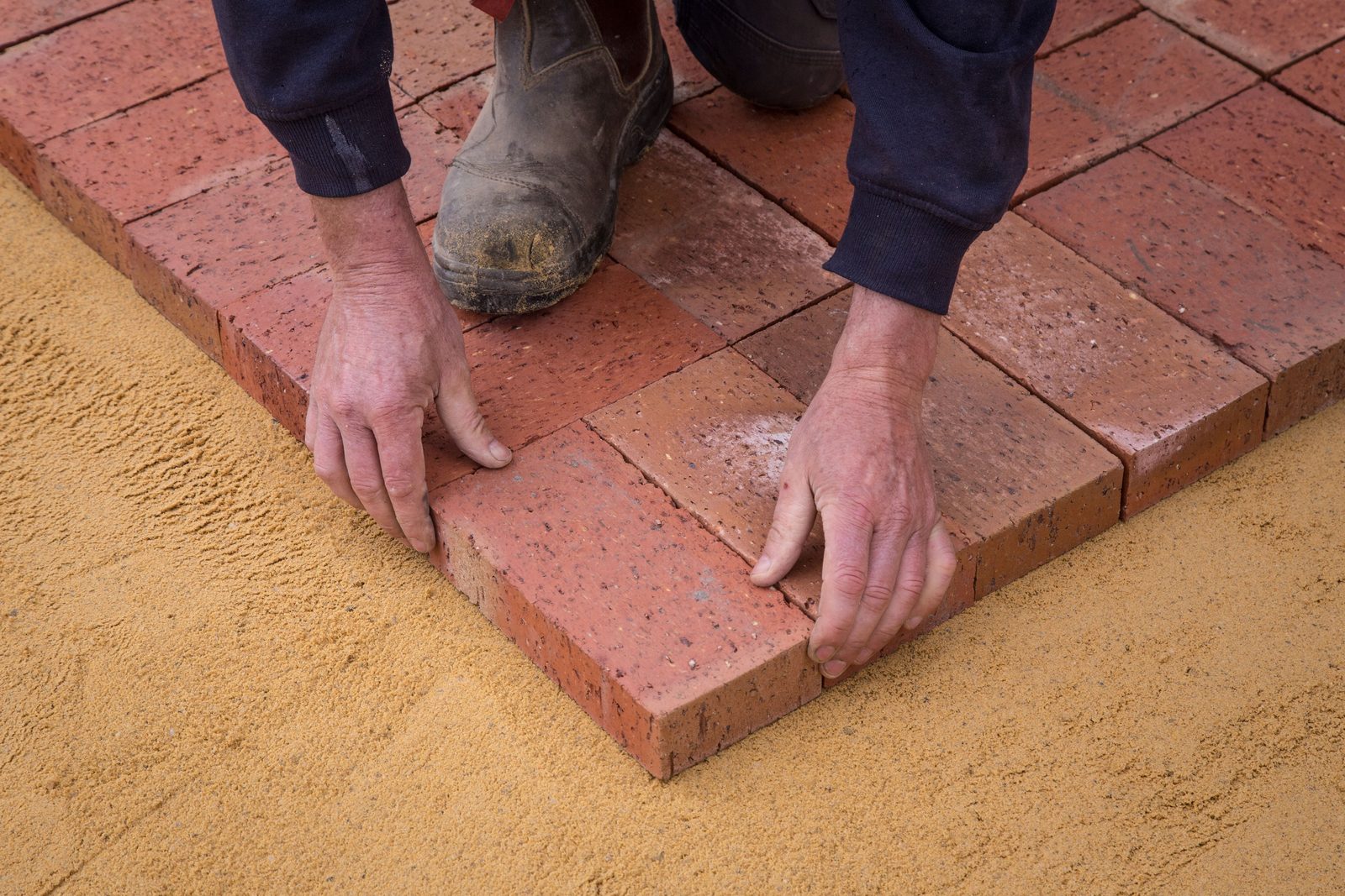 Mason's hands setting red bricks in a row on a sand bed